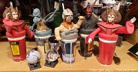 Contents of all seven Star Wars character windup cups with different figures on top. Characters include Queen Amidala, Darth Maul, pilot, droid, and others. The cups have red bodies with Star Wars branding and stand on a flat surface.