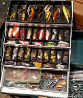 Top view of open tackle box filled with assorted vintage fishing lures organized in compartments, showing various shapes and colors of lures.