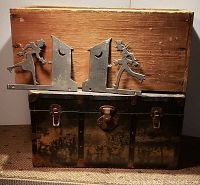 Photo of vintage chest and wooden box with metal outhouse restroom indicator signs placed on top showing rustic condition.