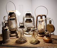 Seven antique and vintage oil burning lamps arranged on woven surface, including metal and glass types with clear chimneys, metal wire handles, some marked Dietz and Beacon.