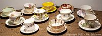 Overview photo showing 11 bone china tea cups with saucers, various floral and patterned designs, arranged on woven mat.