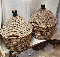 Two large capacity green glass demijohns with conical woven wicker basket exteriors and cork stoppers placed side-by-side on a wooden surface.