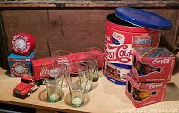 Photo showing Coca-Cola timers boxed, six Coca-Cola glasses, Pepsi-Cola tin, three Coca-Cola boxed mugs, and a small red toy truck.