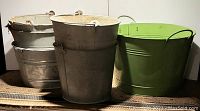 Six water buckets arranged in two rows, five galvanized and one green painted steel bucket visible with metal and wire handles.