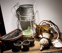 Full view of garden collection items including brass coal scuttle, decorative metal planter on white stand, small black pots, wicker basket, and green ceramic planter.
