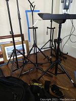 Multiple black metal stands including guitar stands, music stands, microphone stands arranged on wooden floor near wall with framed photos behind.