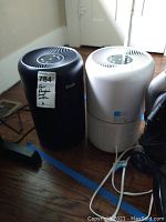 Photo of black and white Levoit Core 300 air purifiers side by side on a wooden floor near a window with sunlight.