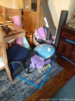 Blue Bosu balance trainer, yoga mats in pink and green rolled up next to basket containing purple therapy balls, foam rollers in various colors, small hand weights, and massage roller under a wooden table on a carpet.