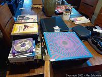 Wooden table with stacks of DVDs and vinyl records including colorful album covers, fitness DVDs, and a DVD audiobook.