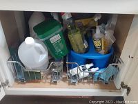 Cleaning supplies organized in two wire racks under a sink, including dish soap, hand soap, bleach bottle, green container, and SOS pads.