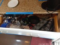 Open kitchen drawer containing pots with lids, a cast iron pan, several stainless steel and metal bowls, and baking trays including muffin tins.