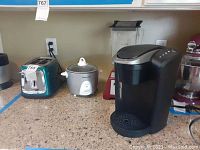 Four kitchen appliances on a countertop: Oster toaster (teal-blue, two-slot), Panasonic rice cooker (white), red Blendtec blender, and black Keurig coffee maker.