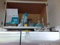 Photo showing upper kitchen cabinet with various items including trash bag boxes, bird seed bag, and glass vases