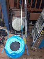 Overall view of two Timber Ridge folding chairs, one chair with plaid back, round side table with cushion, blue collapsible waste basket on wooden deck along with window cleaning tool and ladder nearby