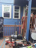 Deck view showing metal round-point shovel, plastic snow shovel, wire-bristle push broom, leaf rake, sledge hammer and cordless string trimmer leaning against railing