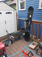 Snowboard and bicycle leaning against deck railing with other items in background.