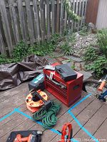 View of red Craftsman toolbox, green and orange extension cords, black plastic Ace socket set case, gloves, and carpet tool on wooden deck