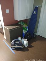 Photo showing laundry hamper, Bissell SpotBot cleaner, iron, fan, large cleaning solution jugs, and packs of paper towels and toilet paper in basement setting.