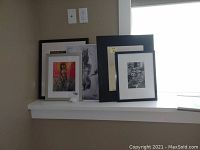 Wide view of multiple framed art prints and mirror leaning on a white ledge near a window.