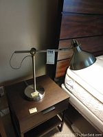 Adjustable chrome finish table lamp on nightstand next to bed. Shows full lamp with conical shade and adjustable arm.