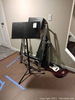 Photo shows a black metal music stand, an electric guitar resting on the floor, a black bass guitar case, a Mexican jazz bass guitar body, and a tripod against a wall.