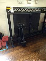 Photo of an iron fireplace screen with decorative scrollwork and four-piece matching tool set including poker, tongs, brush, and shovel, standing on hardwood floor adjacent to fireplace opening.