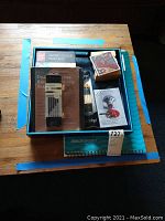 Top-down view of boxed art supplies showing watercolor paint box, drawing pencils in blister package, sketch pad, paint brushes, and art cards on a blue tray.