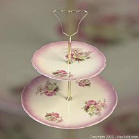 Front view of the two-tiered dessert server showcasing floral decorated plates with pink edge and gold-tone handle.