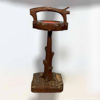 Front angled view of the vintage wooden ashtray stand carved to resemble a tree with branches, square base resembling logs, and top ashtray holder with red insert missing the ashtray itself. The wood is carved in a rustic style with the artist's signature visible on the base.