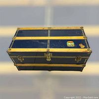 Top view of vintage blue metal and plywood trunk with brass trim and two travel labels, showing worn surface and latch area.