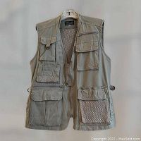 Front view of women's sleeveless outdoor vest in cream. Shows multiple front pockets with zippers and mesh, and side waist adjustment straps.