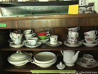 Wide view showing multiple sets of tea cups and saucers, serving dishes, and teapot displayed in wooden cabinet shelf