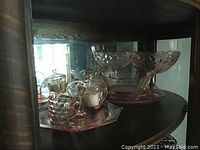 Vintage pink glass items displayed inside a shelf including 2 pedestal candy dishes, creamers, and a handled plate on pink glass trays.