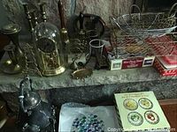 Various brass and metal decorative and kitchen items displayed on a stone hearth, including candlesticks, anniversary clock under glass dome, wire basket, and an assortment of small brass items and a mini pizza stone in its box.