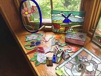 Photo showing multiple stained glass sun catchers on a wooden surface by window light, includes floral designs, bird panel, windmill box, and colored glass orb decorative pieces