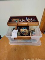 Full view of the wooden tray and plastic bin with assorted jewelry inside including brooches, rings, bracelets, earrings, and a watch