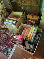 Three boxes of assorted board and card games shown packed with various game boxes inside, visible titles Scrabble, Boggle, checkers, Clue, and more.