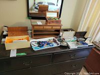Overview of two wooden jewelry boxes on a dark dresser with multiple trays and containers of assorted jewelry items including necklaces, earrings, bracelets, and pins.