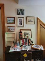 Photo showing multiple framed wall art pieces on the wall and arranged on and above a dark wooden table with various porcelain figurines, books, and a small shelf on top of table.
