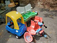 Photo of the set showing the Little Tikes car, red seesaw with green seats, pink-blue tricycle, and a green-red table outdoors on pavement against leaves background.