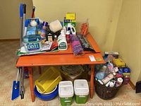 Wide shot of various cleaning products arranged on an orange table and floor showing bottles, brushes, boxes, and assorted supplies.