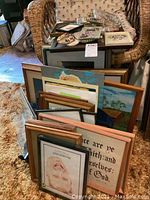 Stack of assorted framed prints and pictures leaning against a couch and a wicker chair, showing different subjects including animals and text.