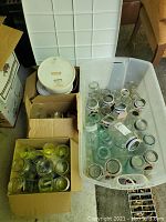 Photo showing multiple glass canning jars in plastic bin and cardboard boxes, some with metal lids, some empty without lids.