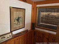 Photo of framed prints on wood-paneled wall, showing one print of a man walking on a path with birds and dog, and a small print of birds below