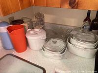 Overview of several kitchen items including multiple plastic pitchers of various colors, Pyrex casserole dishes, a white Tupperware pitcher without lid, and a kitchen scale. Items arranged on a kitchen counter.