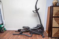 Side view of the elliptical machine showing its black and silver frame, foot pedals and base on wooden floor near a wall.