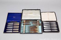 Three vintage boxed flatware sets displayed open showing knives and forks with silver blades and varied handles in blue and white cases