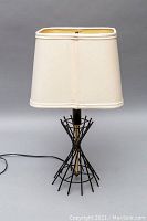 Full front view of the lamp showing metal wireframe base and cream-colored fabric shade, along with attached electrical cord.