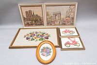 Full view of all six framed needlepoint and art pieces with two cityscape scenes, floral arrangements, and vintage car prints in varied frames.