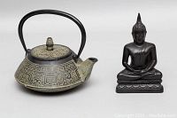 Side view of cast iron teapot with etched pattern alongside small composite Buddha figurine.